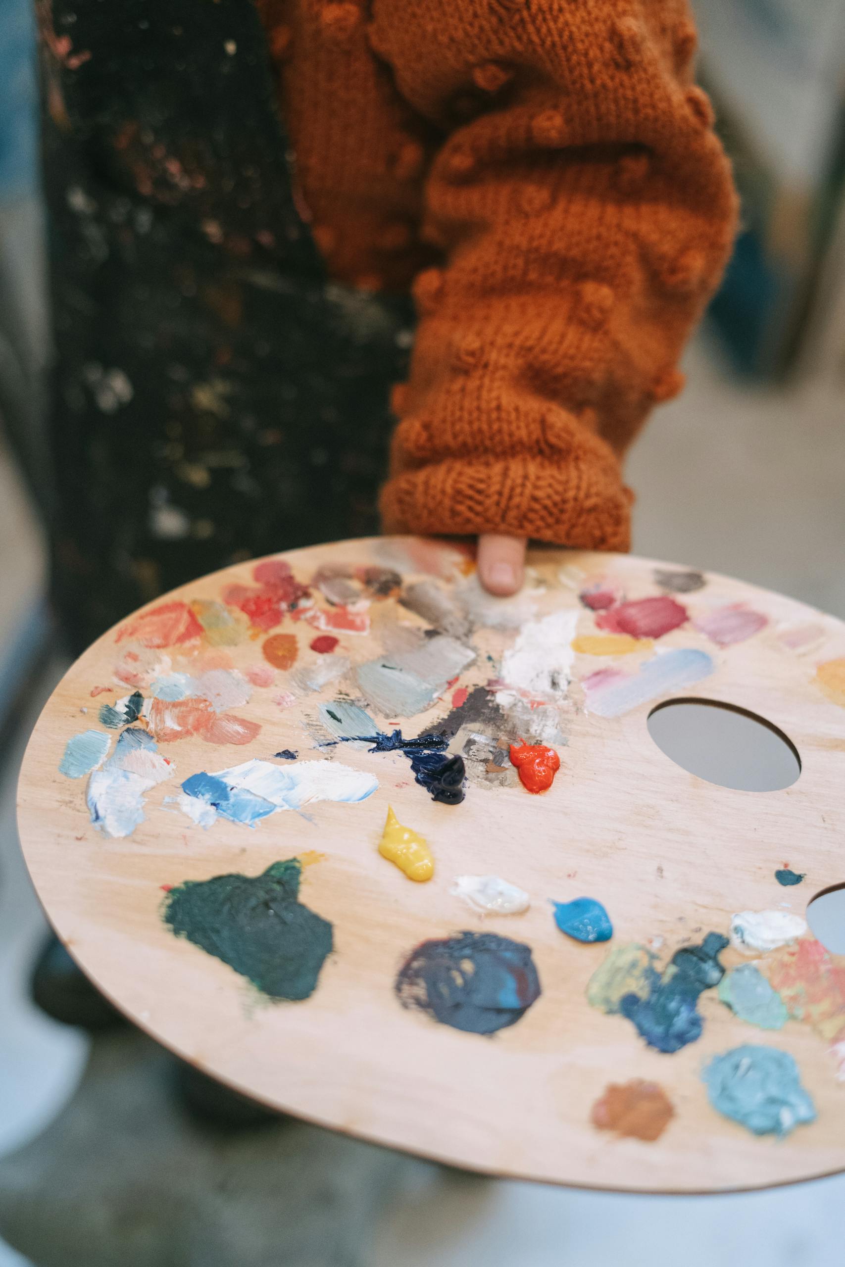 A child in a cozy sweater holding a colorful paint palette during art class.