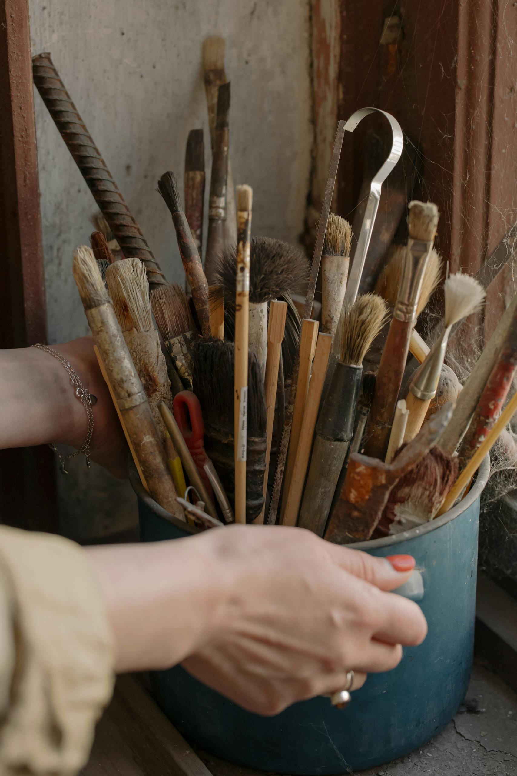 A collection of used paintbrushes in a blue pot by a window, perfect for vintage art themes.