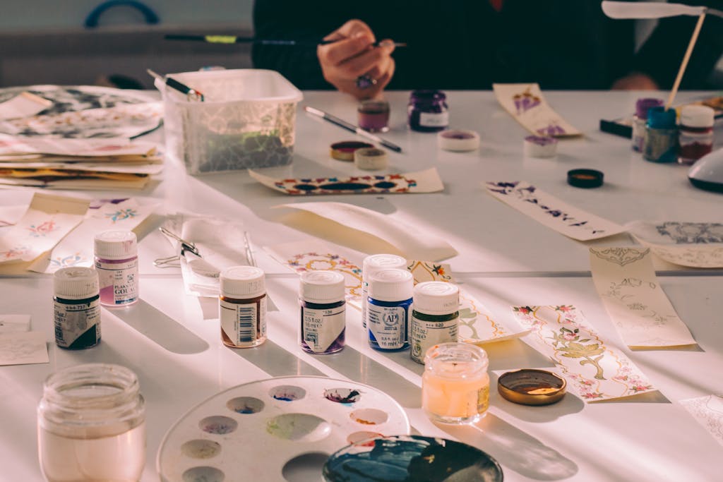 Art studio table with various paints, paper, and an artist's hand crafting designs.