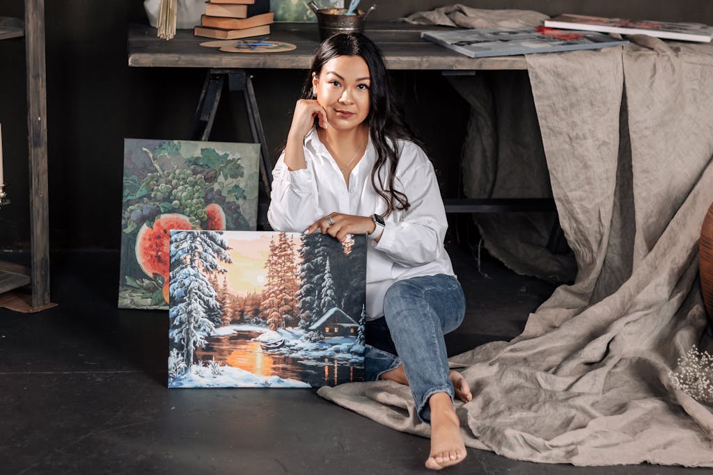 Artist sitting in studio holding a winter-themed painting with a cabin by a lake.
