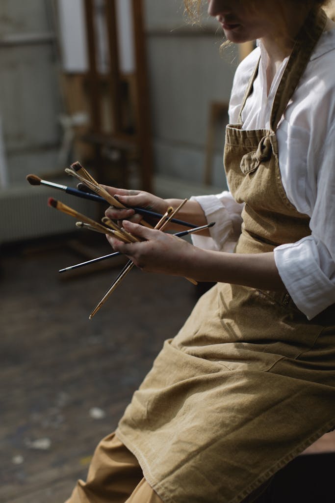 Artist with apron holding multiple paintbrushes in a dimly lit studio environment, highlighting creativity.
