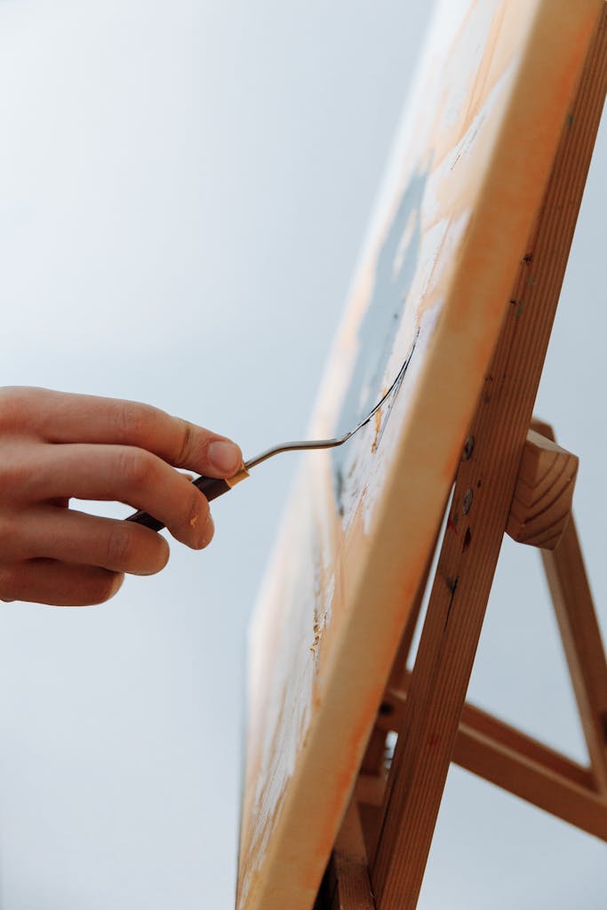 Close-up of an artist's hand painting with a palette knife on a canvas on an easel.