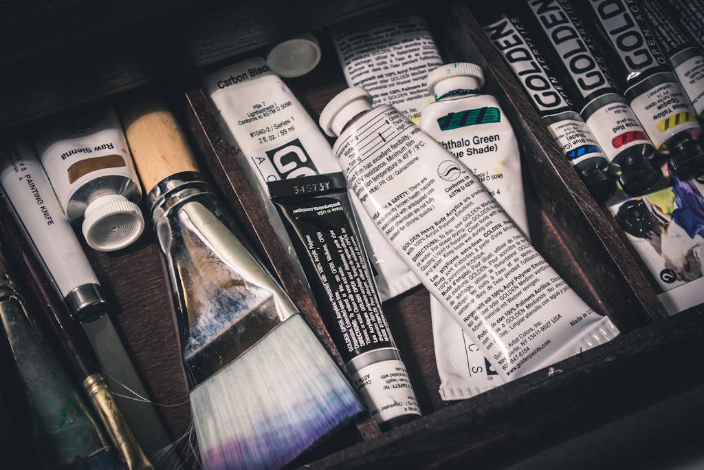 Close-up of art supplies including paint tubes and brushes arranged in a wooden box.