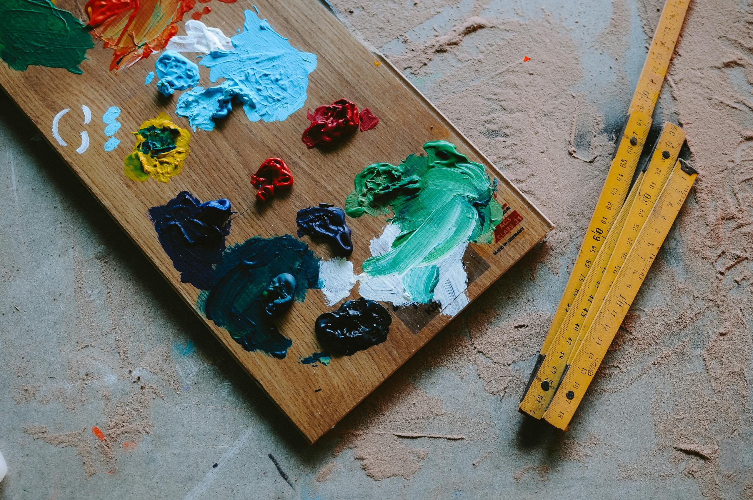 Colorful oil paints on a wooden palette beside wooden measuring tapes on a textured floor.
